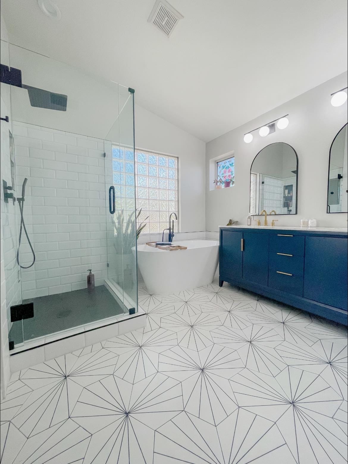 Gilbert master bathroom after — navy blue freestanding double vanity with arched mirrors, subway tile walk-in shower, freestanding soaking tub, and graphic starburst tile floor