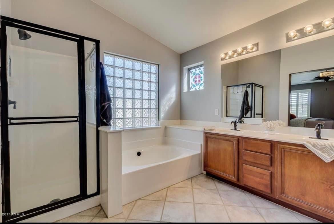 Gilbert second-story master bathroom before — original cherry double vanity, garden tub under glass-block windows, and black-framed shower stall