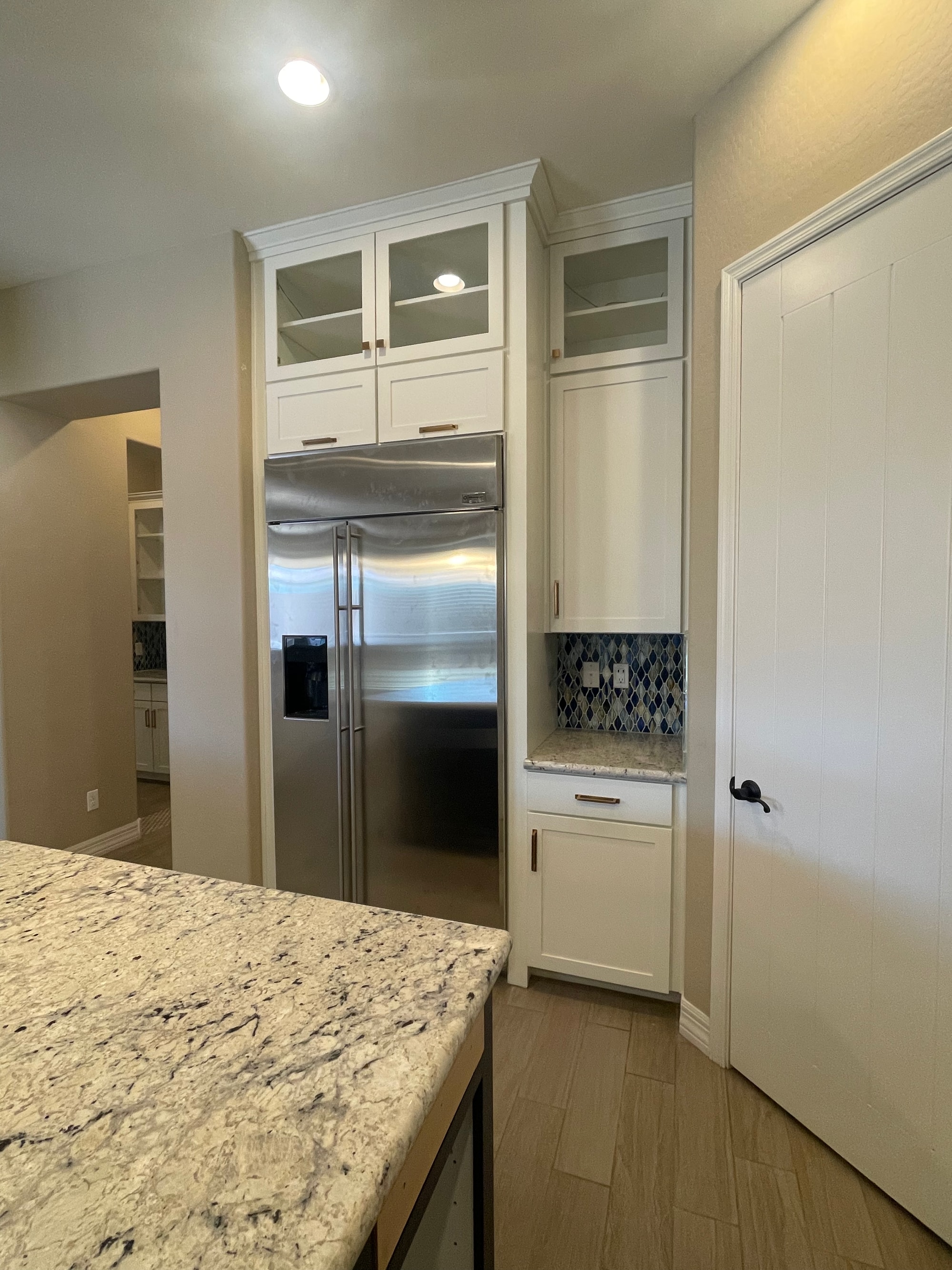 Queen Creek kitchen — corner over the built-in stainless refrigerator showing the new glass display cabinets extended to the ceiling, gold pulls, and existing granite countertop and backsplash preserved on the side counter